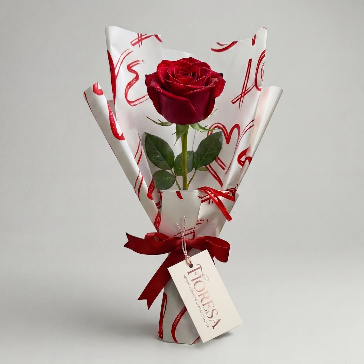 Fioresa single red rose stem elegantly wrapped in white love-print paper with red ribbon bow and branded gift tag, perfect romantic gifting in Surat.