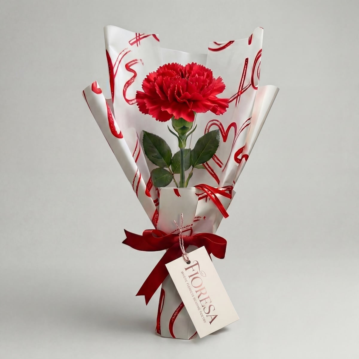 A single red carnation flower in a Fioresa heart-themed gift wrap with a red ribbon and branded tag on a clean white background.