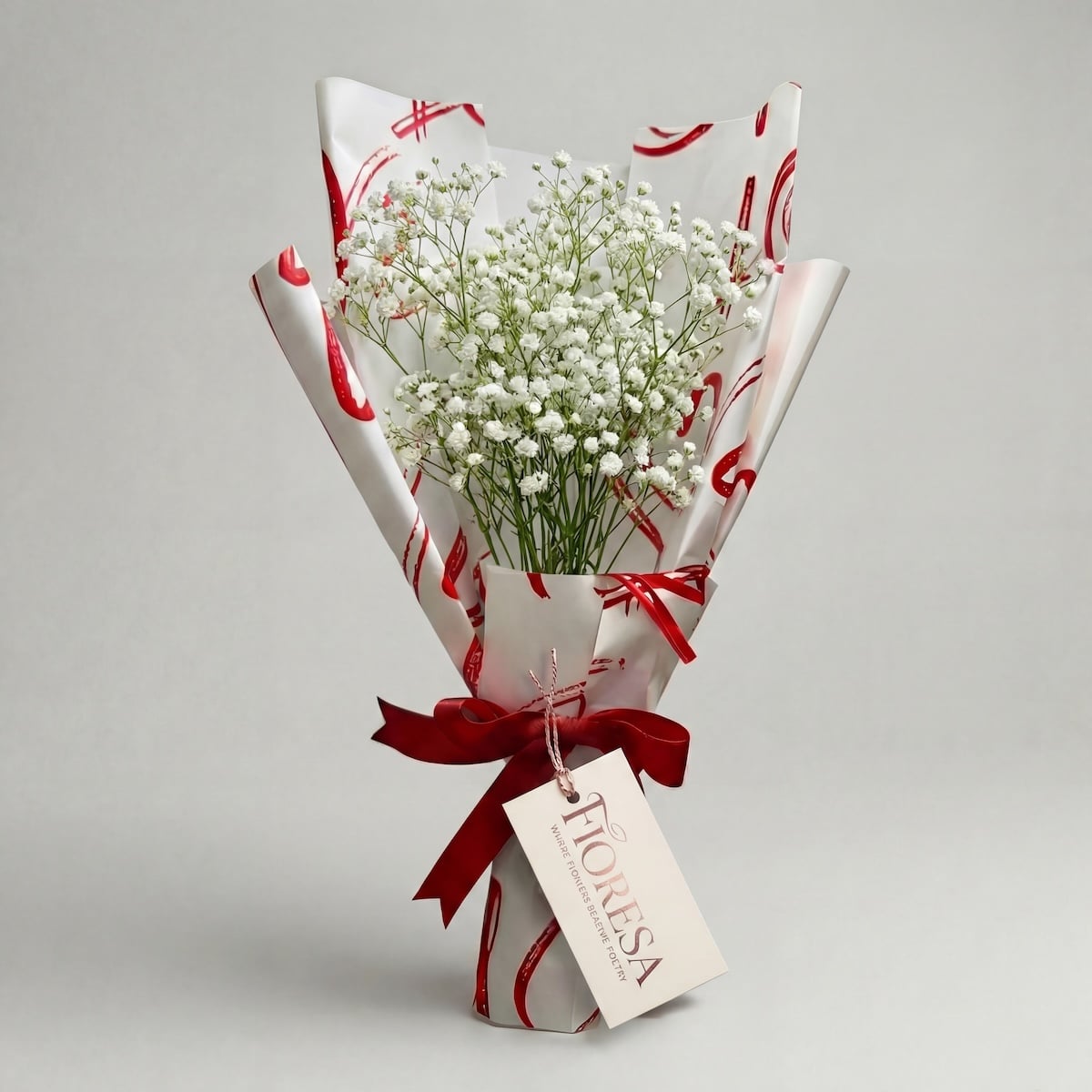 A delicate bouquet of white baby's breath (gypsophila) in a Fioresa heart-patterned gift wrap with a red ribbon and branded tag on a neutral background.