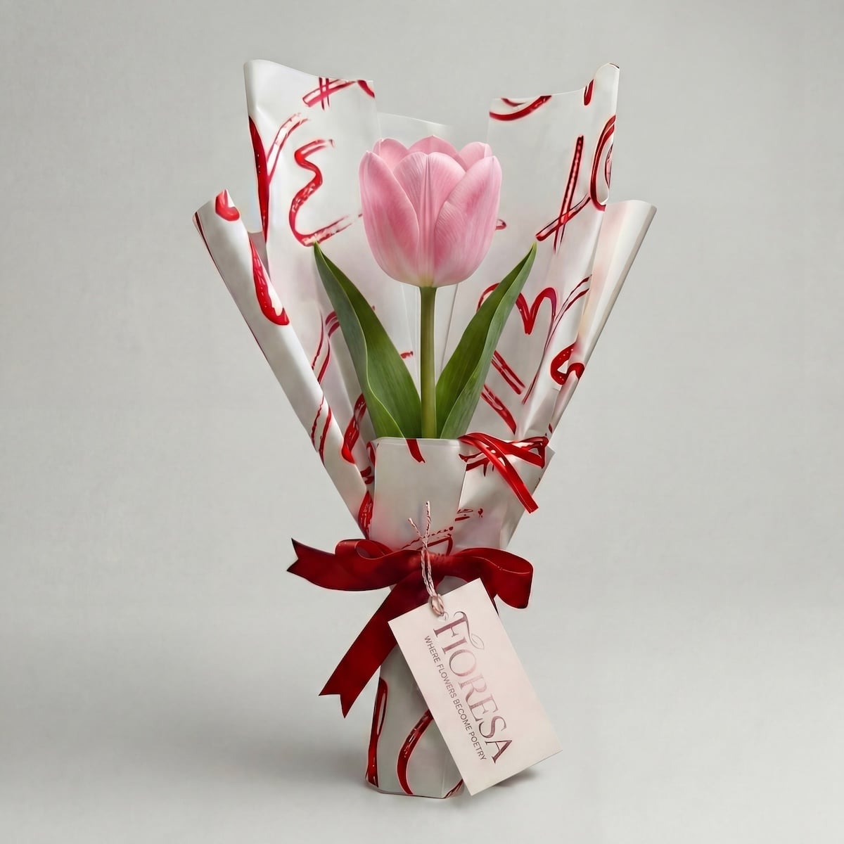 Fioresa single pink tulip stem wrapped in romantic white love-print paper with red ribbon and branded tag, elegant luxury flower gift from Surat.