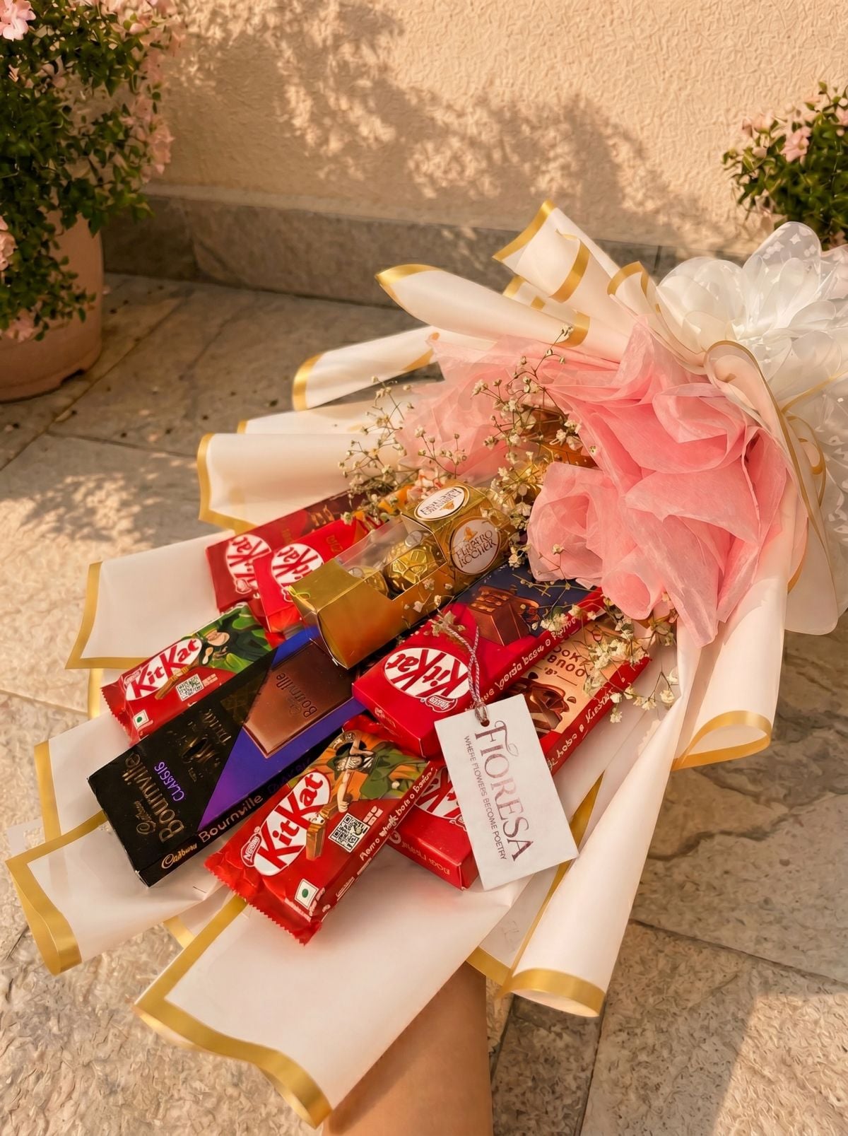 A chocolate gift bouquet from Fioresa arranged horizontally. The bouquet features various chocolates including multiple red KitKat bars, a purple and black Cadbury Bournville Classic dark chocolate bar, and a small gold box of Ferrero Rocher pralines. The chocolates are decorated with delicate white baby's breath flowers and light pink ruffled tissue paper. The arrangement is wrapped in premium white paper with gold edges and tied with a large white translucent bow at the top right. A white brand tag attached to the center reads "FIORESA — WHERE FLOWERS BECOME POETRY." The bouquet rests against a textured tiled ground with green potted plants visible in the background.